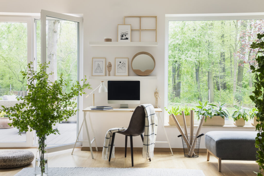 Real photo of white living room interior with big window, glass door, fresh plants, wooden desk with mockup computer and simple posters on shelves