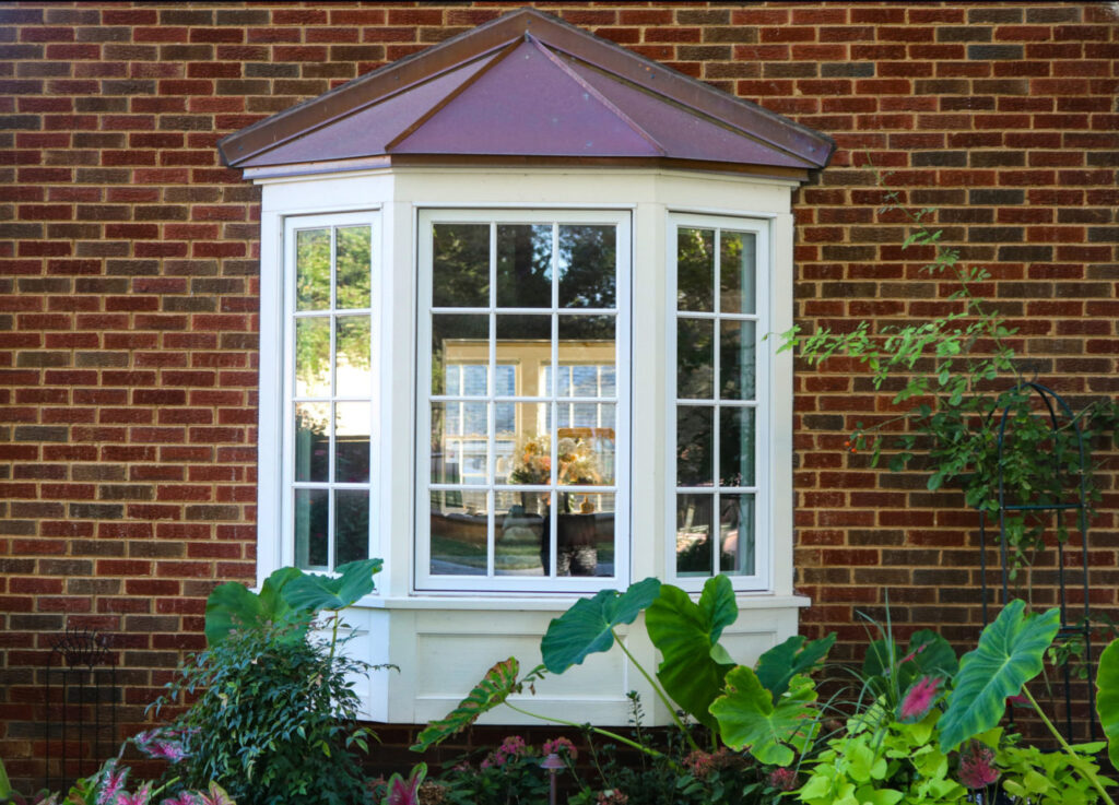 Bay window in a brick house with reflection of trees and view of windows and flowers inside and flowers