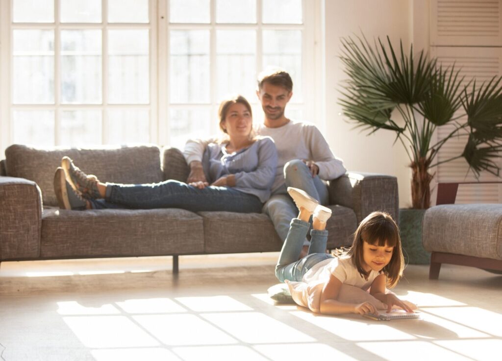 Happy Family lounges on couch with sunlight streaming in while daughter draws on the carpet 