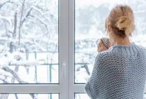 A woman in a cozy sweater stands at the picture window with a cup of drink in her hands